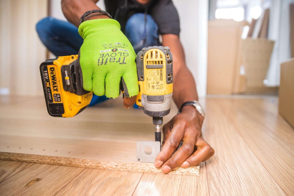 A man with a drill in his hand, wearing a light green glove, is drilling a hole in a piece of light brown wood that is on the floor.