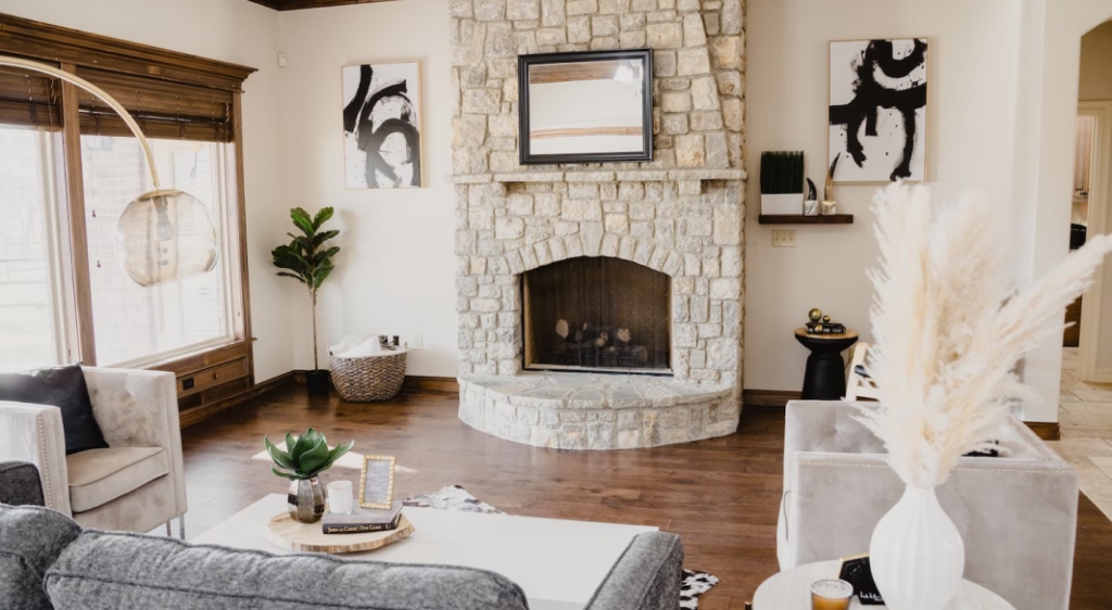 A living room with white and beige colors, with a fireplace at the center of the room, painting on the calls, plants here and there, a light fixture and a wooden floor.